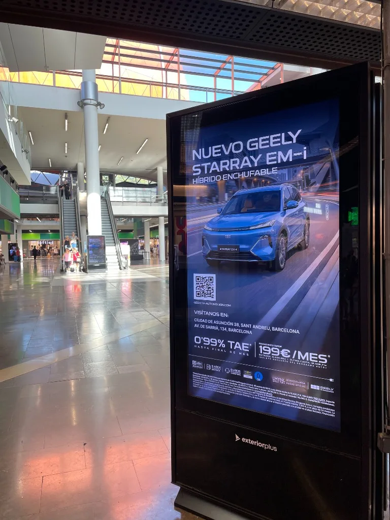 Tòtem digital publicitari vertical situat al costat de les escales mecàniques d’un centre comercial